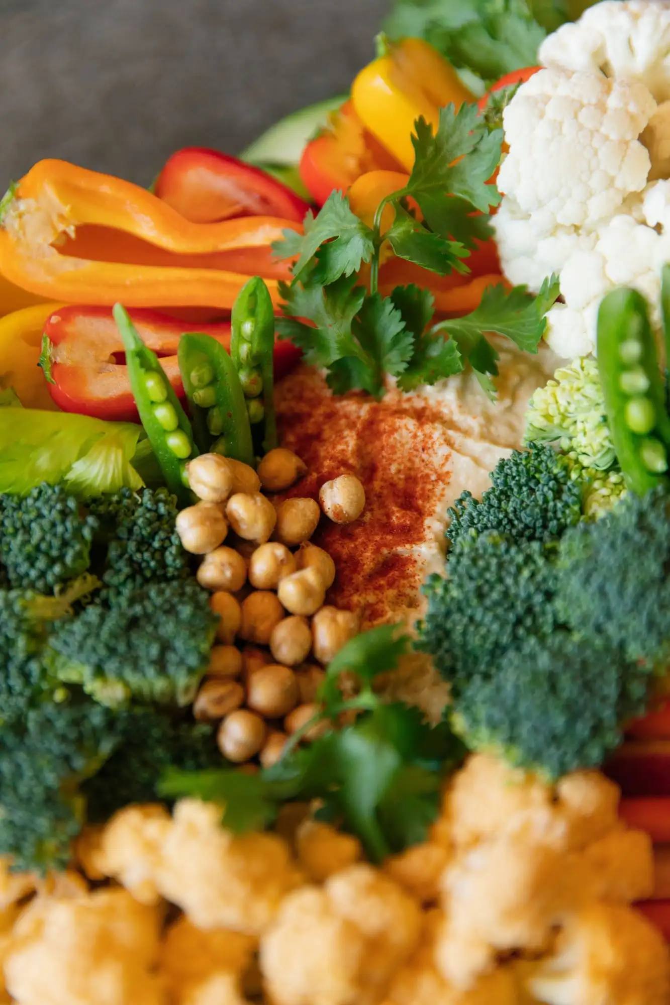 Board of rainbow carrots, peppers, snap peas and tomatoes around a bowl of hummus (view 4)