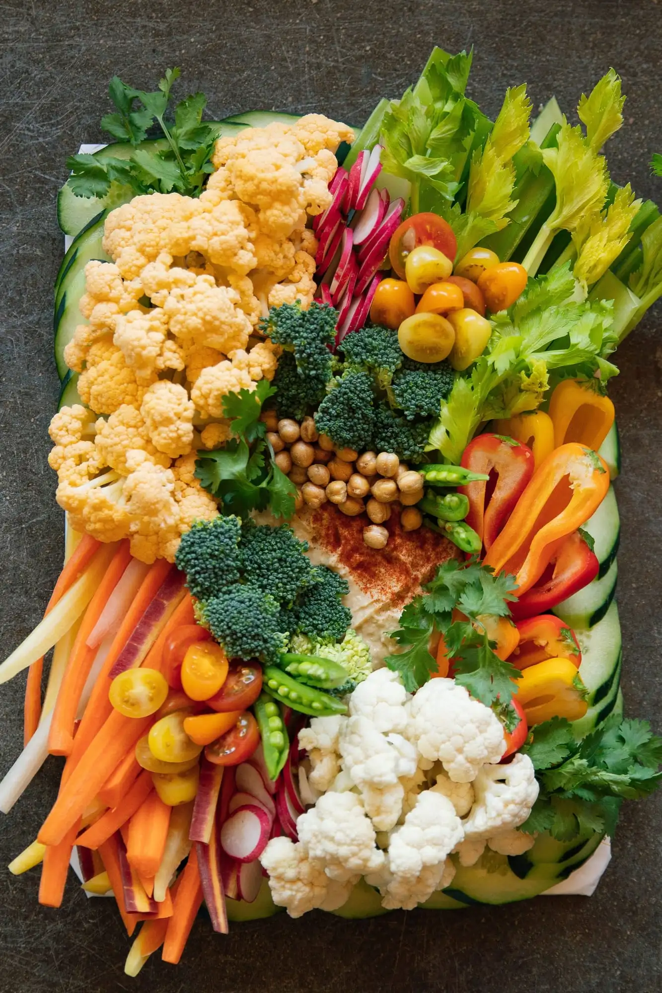 Board of rainbow carrots, peppers, snap peas and tomatoes around a bowl of hummus