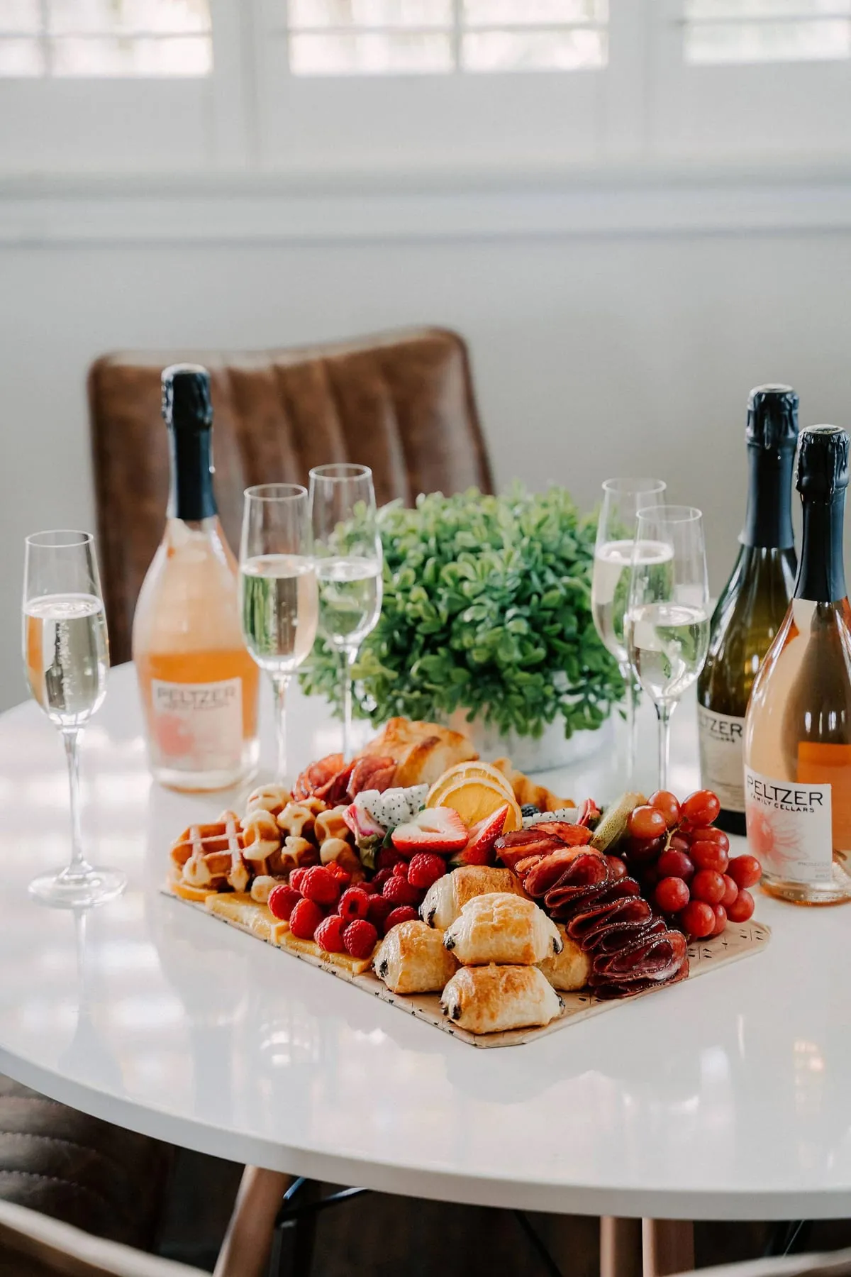 A brunch board with croissants, berries and waffles beside bottles of sparkling wine