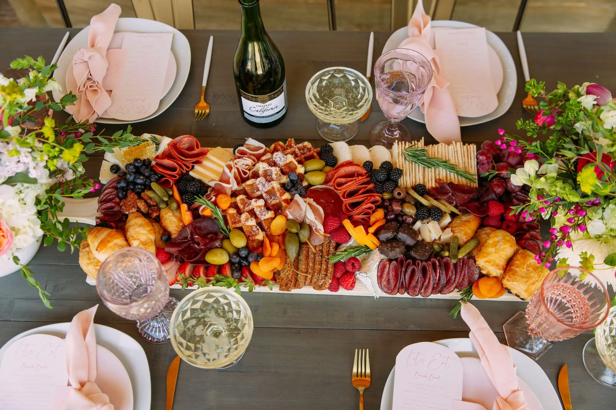 A long brunch grazing board of waffles, croissants, charcuterie, cheeses and berries on a table set with pink napkins, flowers and sparkling wine
