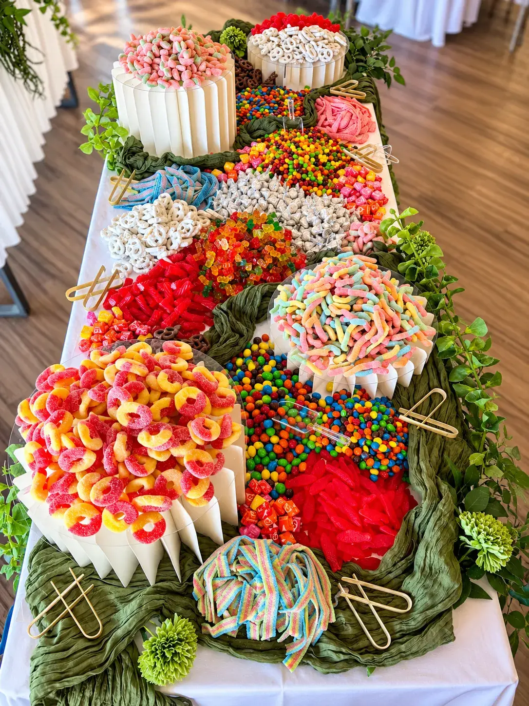 Our candy grazing table for 100 guests at the Visit Temecula Valley Marketing Summit: peach rings, sour worms, gummy bears and chocolates on white risers with green runners and greenery