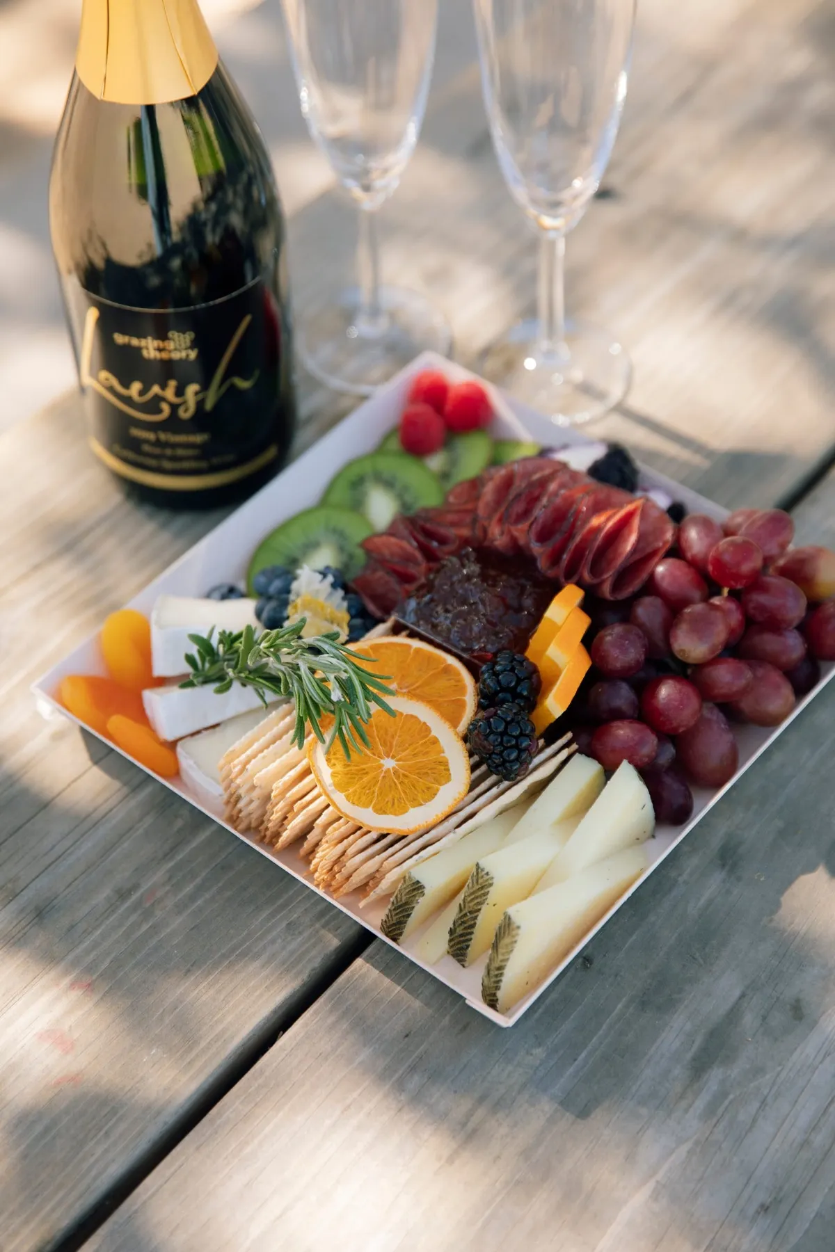 A cheese and charcuterie board with salami, oranges, grapes and crackers on a picnic table