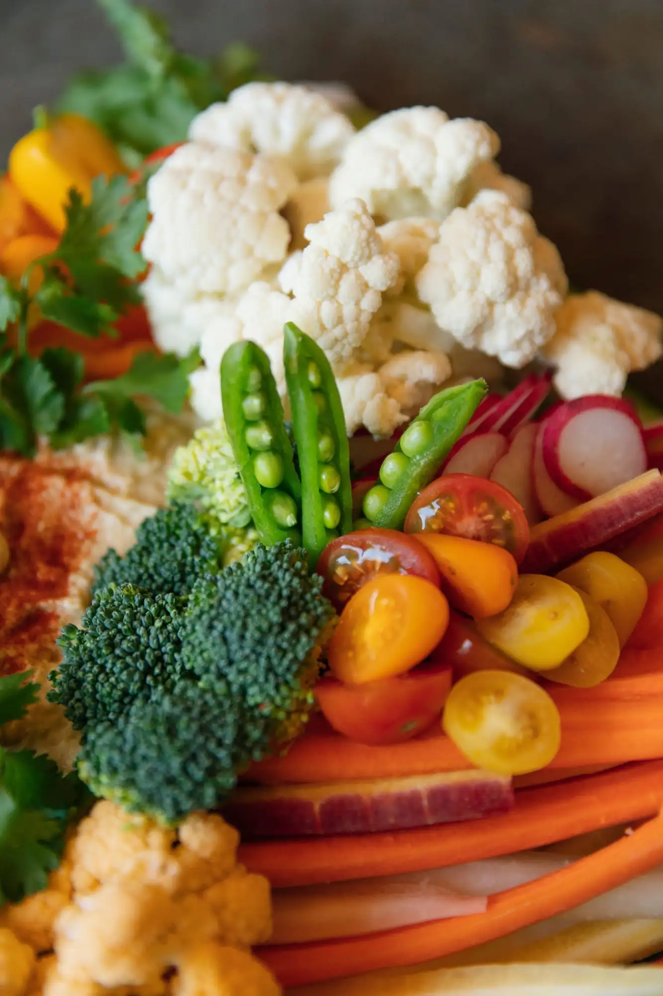 Board of rainbow carrots, peppers, snap peas and tomatoes around a bowl of hummus (view 3)