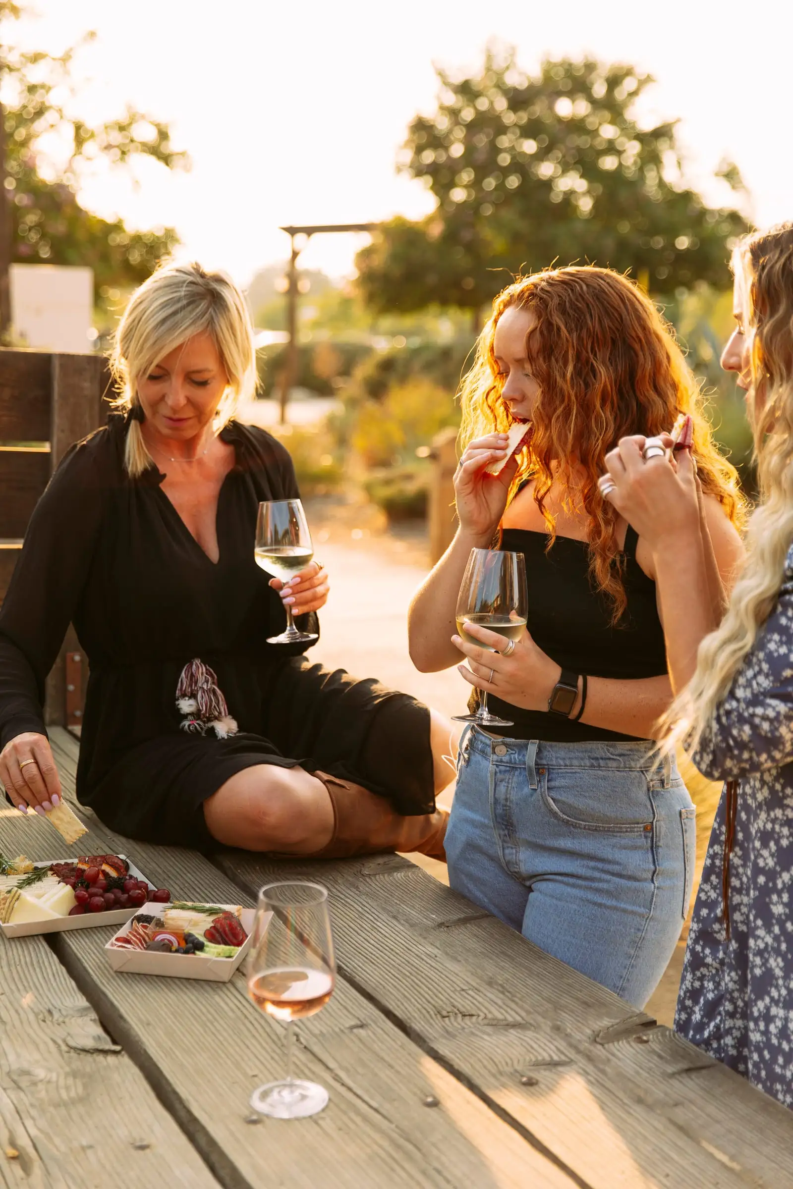 Friends sharing charcuterie boxes and wine at an outdoor table at golden hour