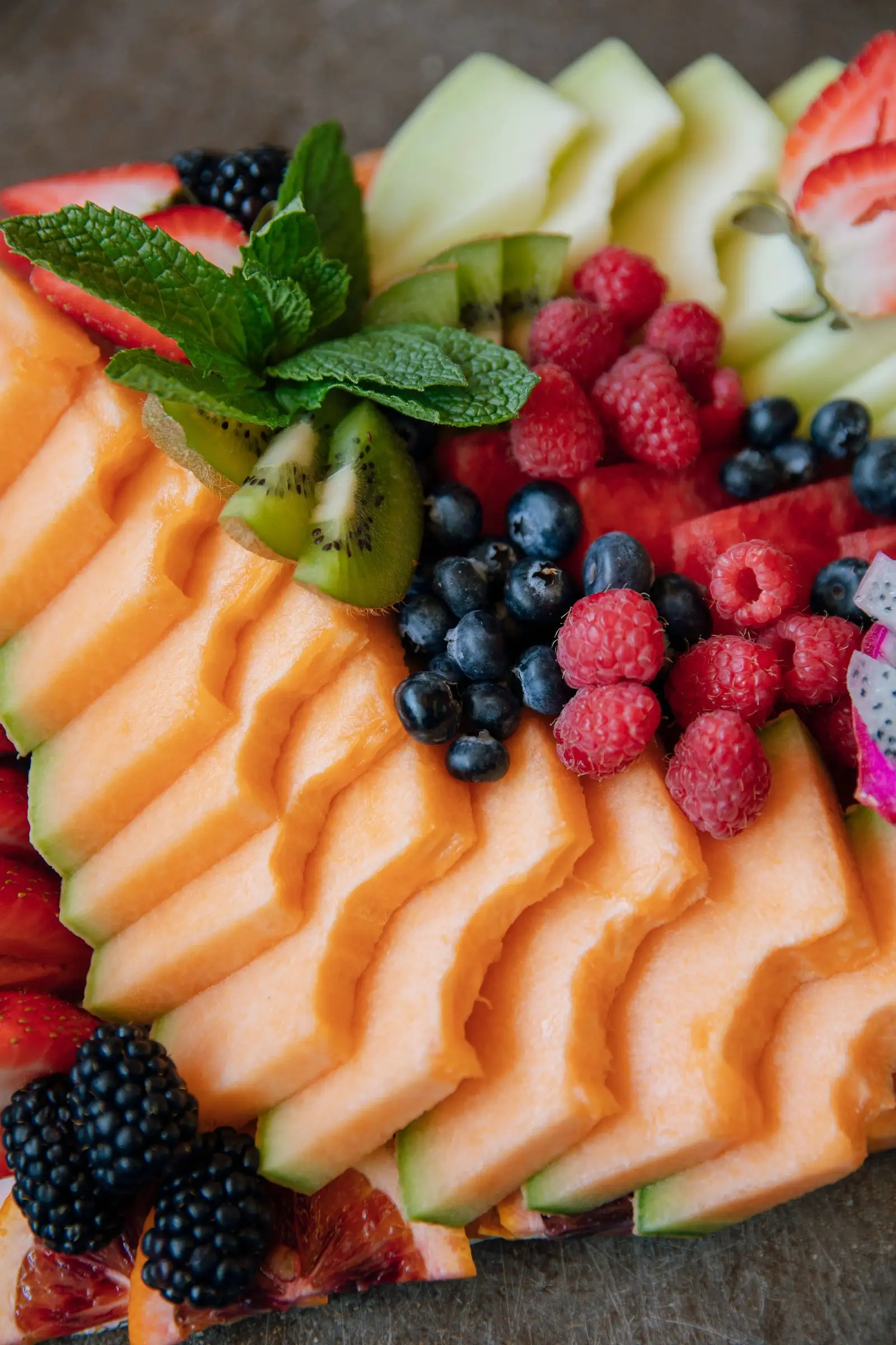 A fruit board of fanned cantaloupe and honeydew with raspberries, blueberries, kiwi, blackberries and fresh mint