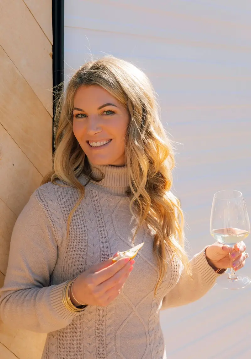 Gina LaMora smiling, holding a glass of white wine and a bite of cheese