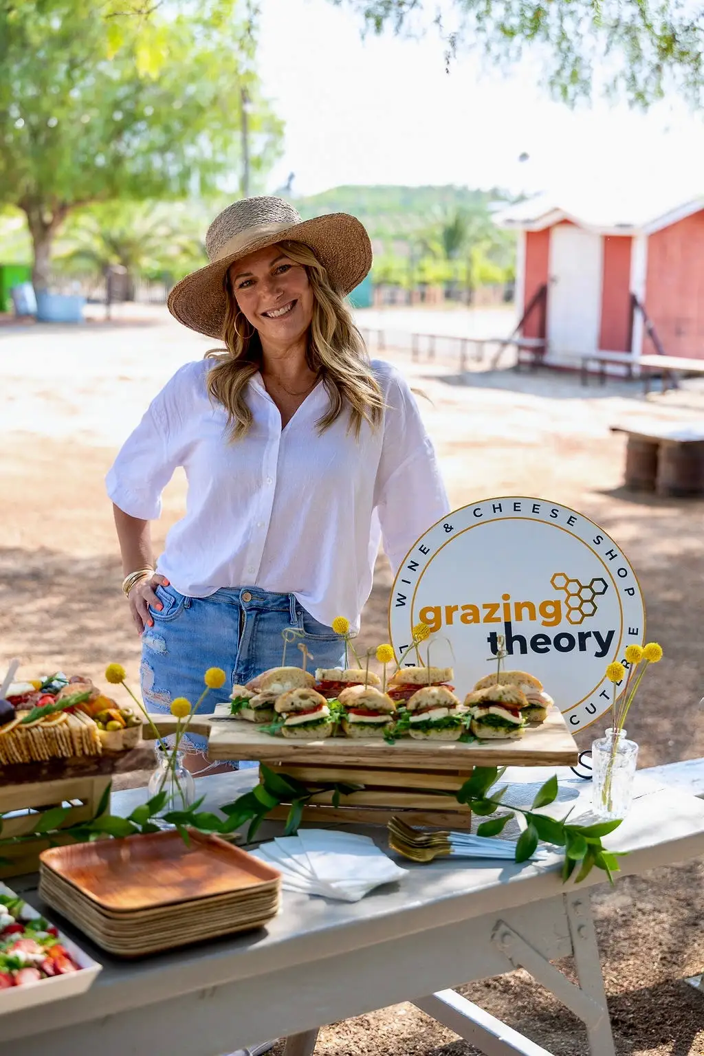 Gina LaMora in a sun hat beside an outdoor grazing table of sandwiches, charcuterie and fresh flowers