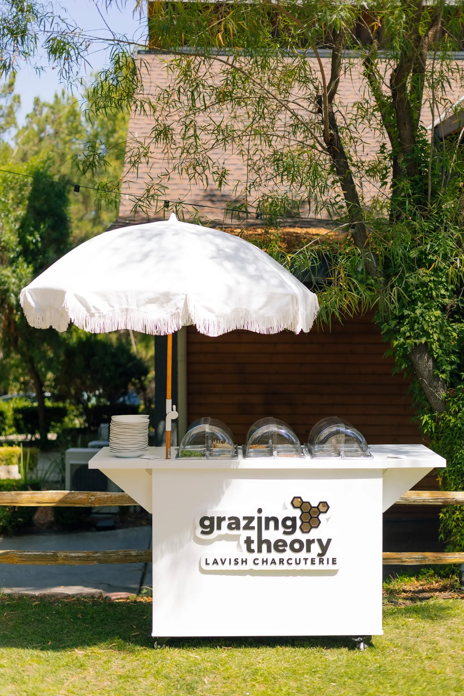 The Grazing Theory mobile charcuterie cart under a white fringed umbrella at a garden event