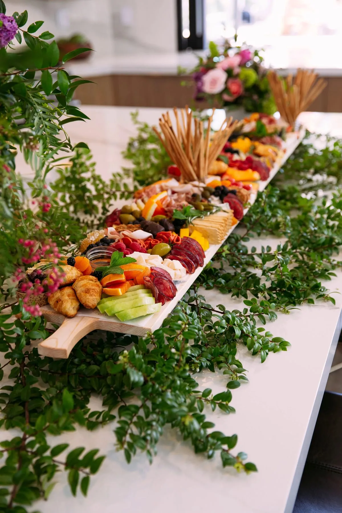 A multi-level grazing table with greenery, cheeses, fruit and breadsticks
