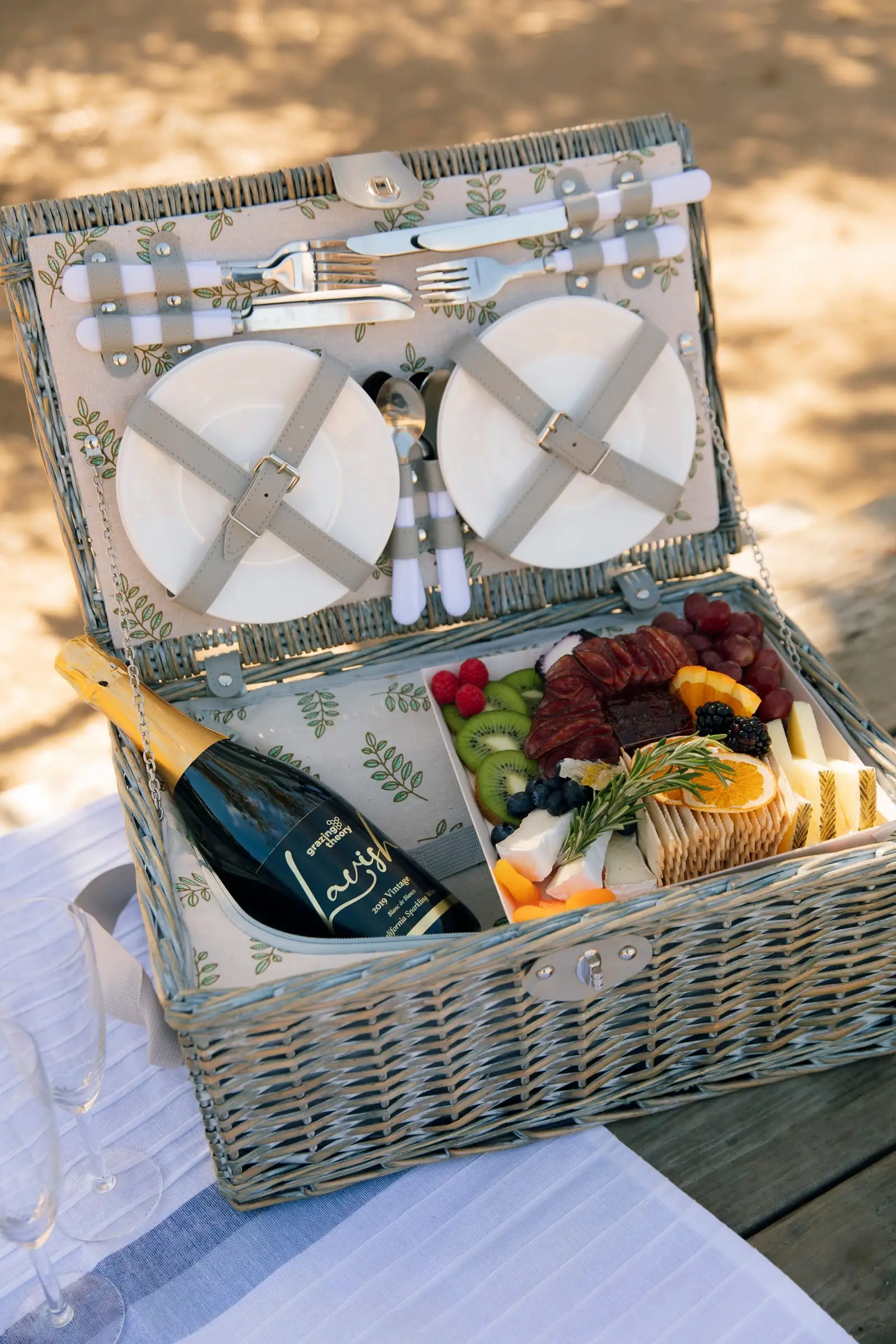 A wicker picnic basket with plates, a bottle of sparkling wine and a charcuterie board on a wooden picnic table