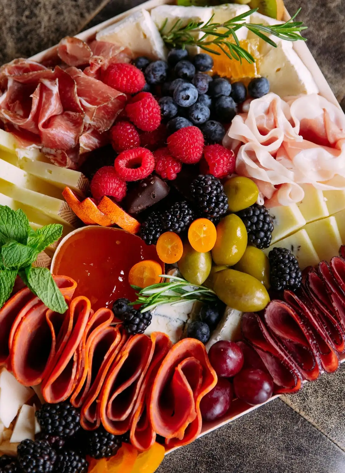 A charcuterie board with prosciutto, folded salami, cheeses, olives, berries, kumquats and a cup of jelly