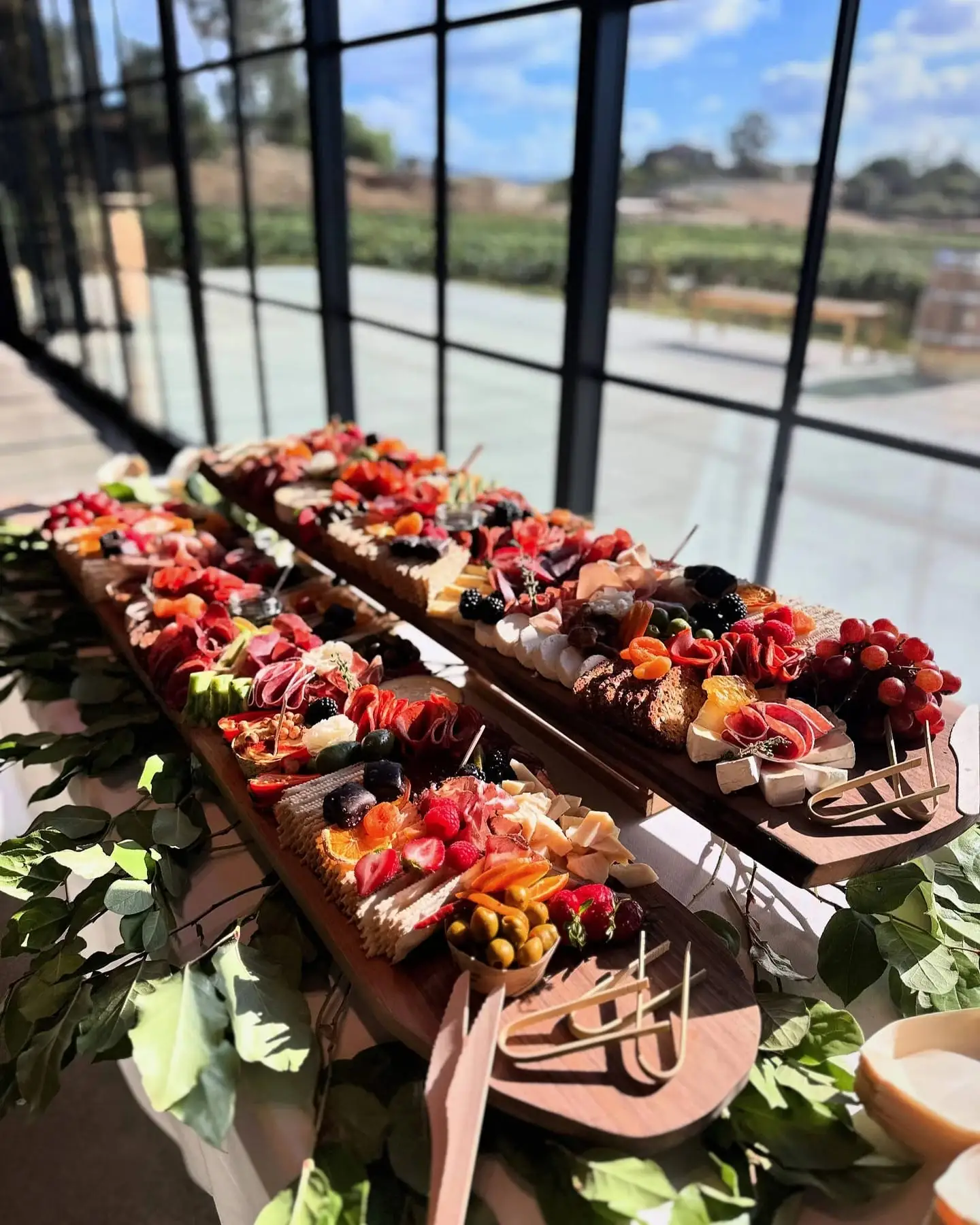 Long wedding grazing tables of cheeses, cured meats and fruit beside tall windows overlooking Temecula vineyards